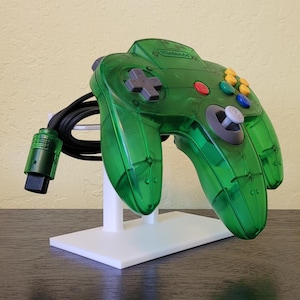 Könnte beinhalten: Ein grüner, durchsichtiger Nintendo 64-Controller mit einem weißen Ständer. Der Controller hat einen grauen Analogstick und gelbe, blaue und rote Knöpfe.