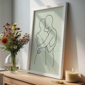 May include: A framed minimalist line art print featuring two embracing figures in black against a light green background. The artwork is displayed on a wooden table, alongside a vase of flowers and a lit candle. The text at the bottom reads: "Minimalist line art - timeless and elegant."