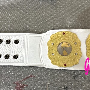 Op de afbeelding: Een witte worstelkampioensriem met goudkleurige medaillons en rode accenten. De riem heeft een gedetailleerd ontwerp met een wereldkaart op de centrale medaillons. De tekst "Grid Gra Sports" is zichtbaar.
