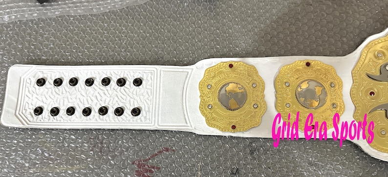 Op de afbeelding: Een witte worstelkampioensriem met goudkleurige medaillons en rode accenten. De riem heeft een gedetailleerd ontwerp met een wereldkaart op de centrale medaillons. De tekst "Grid Gra Sports" is zichtbaar.