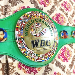 May include: A green leather championship belt featuring a large circular gold center plate with the text WORLD CHAMPION and WBC. The plate is bordered by small international flags. Three circular portraits are attached to the green strap.