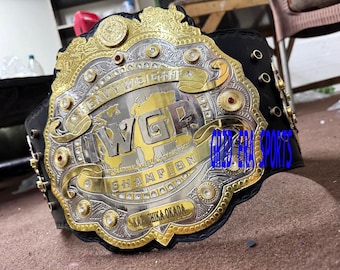 Pode incluir: Um cinto de campeonato com detalhes dourados e prateados, com o texto "HEAVY WEIGHT" e "IWGP INTERNATIONAL WRESTLING GRAND PRIX CHAMPION". O cinto tem uma correia de couro preta e é adornado com pedras preciosas vermelhas e transparentes.