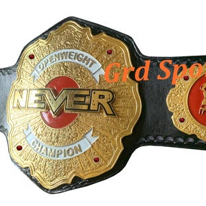 Puede incluir: Un cinturón de campeonato de cuero negro con una gran placa central dorada con el texto NEVER OPENWEIGHT CHAMPION, una incrustación circular roja y placas laterales doradas más pequeñas. El texto Grd Sports se ve en naranja en la correa.