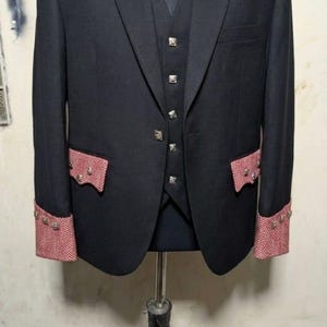 May include: A black suit with a matching vest, displayed on a mannequin. The jacket features red and white patterned accents on the cuffs and pockets, with decorative silver buttons. The shoulders have a red and white trim.