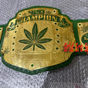 Puede incluir: Un cinturón de campeonato dorado y verde con el texto "420 CHAMPION". El cinturón presenta una gran hoja de cannabis en el centro y hojas más pequeñas en las placas laterales. El cinturón es de metal y tiene una correa verde.