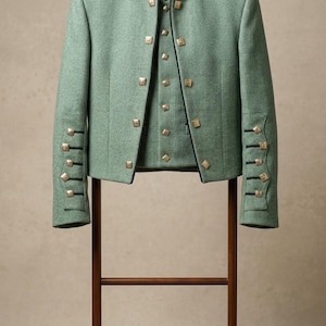 Puede incluir: Chaqueta verde claro con botones cuadrados dorados y ribetes negros, exhibida en una percha de madera. La chaqueta tiene un cuello negro y botones decorativos en las mangas. La chaqueta mide aproximadamente 76 cm de largo.
