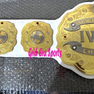 Op de afbeelding: Een gouden worstelkampioenschapsriem met de tekst "INTERCONTINENTAL IWC CHAMPION". De riem heeft een witte band en drie gouden medaillons met een wereldbolontwerp en rode accenten.