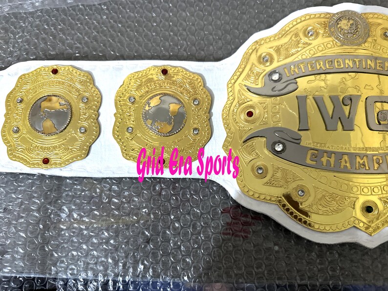 Op de afbeelding: Een gouden worstelkampioenschapsriem met de tekst "INTERCONTINENTAL IWC CHAMPION". De riem heeft een witte band en drie gouden medaillons met een wereldbolontwerp en rode accenten.