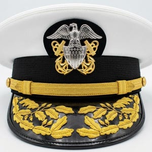 Könnte beinhalten: Weißer Marineoffiziershut mit schwarzem Band und goldenem Visier. Der Hut hat ein goldenes Emblem mit einem Adler und einem Schild sowie eine Goldblattstickerei auf dem Visier. Ein formelles militärisches Accessoire.
