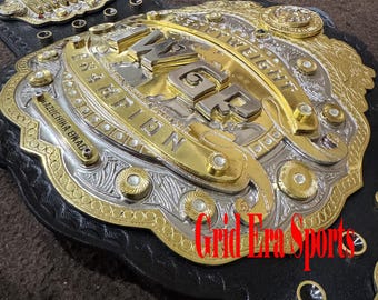 May include: A close-up of a wrestling championship belt. The belt features gold and silver metal detailing with the words "Heavyweight Champion" and "IWGP" visible. The belt is black leather.