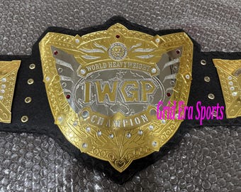 May include: A gold and silver wrestling championship belt with the text "WORLD HEAVYWEIGHT IWGP CHAMPION". The belt has a black leather strap and gold side plates with a globe design. Red jewels are set into the main plate.