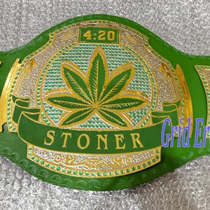 Puede incluir: Cinturón de campeonato verde y dorado con un diseño de hoja de cannabis. El cinturón presenta el texto "STONER" en una pancarta, con "4:20" encima de la hoja. Es un artículo deportivo.