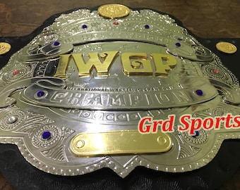 May include: A championship wrestling belt featuring a large silver-toned metal plate with gold-toned lettering that reads IWGP CHAMPION and INTERNATIONAL WRESTLING GRAND PRIX. The plate is adorned with blue and red rhinestones and the text Grd Sports.