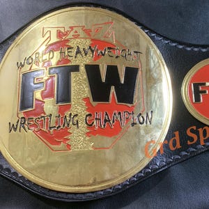 May include: A black leather championship belt featuring a large circular gold-tone center plate with the text TAZ WORLD HEAVYWEIGHT FTW WRESTLING CHAMPION in black and red. The strap includes smaller circular side plates with red centers and FTW text.