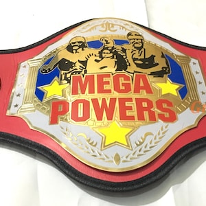 May include: A wrestling championship belt with a red leather strap and gold-colored metal accents. The center plate reads "MEGA POWERS" in red lettering, with images of wrestlers. The side plates say "MACHO MADNESS".