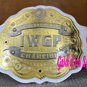 Op de afbeelding: Een gouden en zilveren worstelkampioenschapsriem met de tekst "INTERCONTINENTAL I.W.G.P. CHAMPION" op een witte leren band. De riem heeft ingewikkelde details en decoratieve elementen.