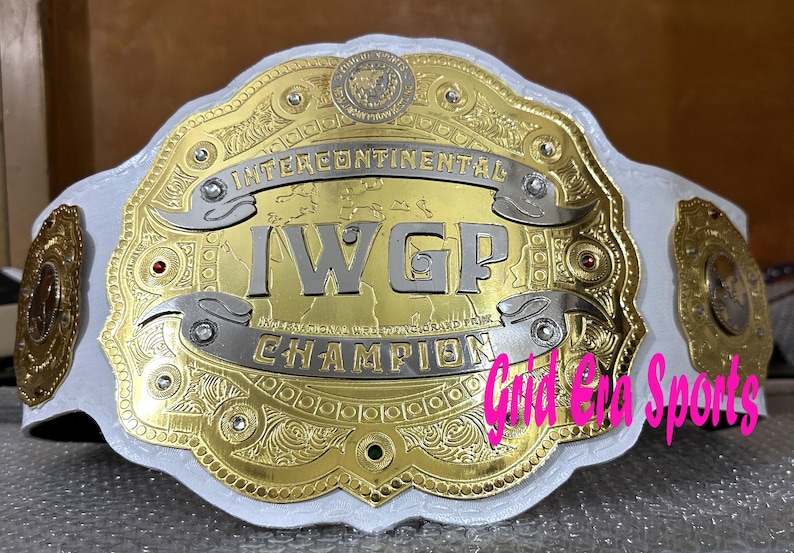 Op de afbeelding: Een gouden en zilveren worstelkampioenschapsriem met de tekst "INTERCONTINENTAL I.W.G.P. CHAMPION" op een witte leren band. De riem heeft ingewikkelde details en decoratieve elementen.