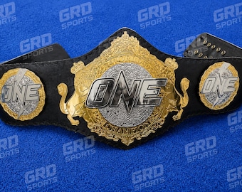 Puede incluir: Un cinturón de campeonato de cuero negro con una gran placa central dorada y plateada con el texto ONE WORLD CHAMPION, flanqueado por dos placas circulares más pequeñas doradas y plateadas que muestran el texto ONE con un rayo.