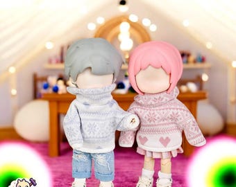 May include: Two small dolls wearing winter sweaters. The doll on the left is wearing a blue and white sweater and blue jeans. The doll on the right is wearing a pink and white sweater and a pink skirt.