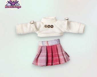 May include: A white doll top with long sleeves and a plaid skirt. The top has a turtleneck and three gold buttons. The skirt is red and white plaid with pleats.