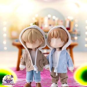 May include: Two dolls wearing hooded jackets. One doll is wearing a brown jacket with a white hood and the other doll is wearing a light blue jacket with a white hood. Both dolls are wearing jeans and have brown hair.