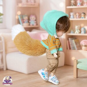 May include: A doll with brown hair wearing a mint green hooded sweatshirt, a mustard and brown striped long-sleeve shirt, and tan cargo trousers. The doll features a large, fluffy, tan and white fox tail and wears white trainers with iridescent accents.