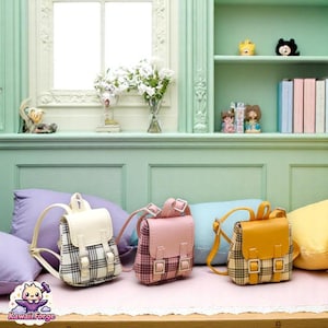 Puede incluir: Tres mini mochilas en crema, rosa y amarillo con estampados de cuadros. Cada una tiene un cierre de solapa y correas ajustables. Las mochilas se exhiben sobre una superficie rosa con almohadas en el fondo.