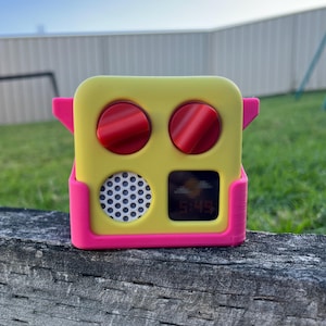 May include: A yellow and pink digital clock with two red knobs, a speaker, and a digital display showing the time. The clock is on a wooden surface, with a green grassy background.
