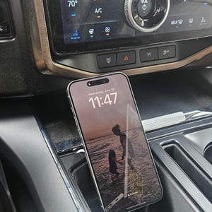 May include: A smartphone with a beach scene wallpaper rests in a car's console. The screen displays "Wednesday, June 18, 11:47." The car's dashboard features climate controls, a volume knob, and a display showing "This Trip 1.8 mi/kWh."