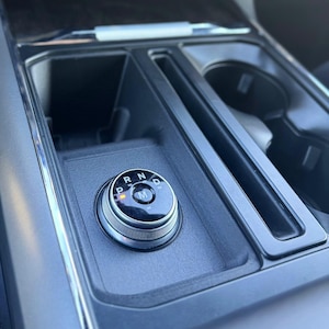 May include: Close-up of a car's center console featuring a rotary gear selector. The selector is a silver-colored dial with the letters P, R, N, and D, and an M in the center. The console is dark gray with cup holders and storage compartments.