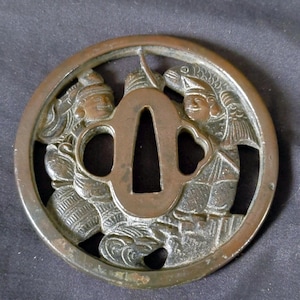 Op de afbeelding: Een ronde bronzen tsuba met een opengewerkt ontwerp van twee figuren in traditionele kleding. Het centrale gedeelte bevat een driehoekige opening voor een zwaardblad en twee kleinere zijopeningen, omringd door gedetailleerde reliëfs.