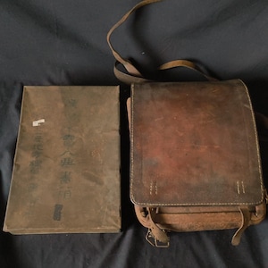 May include: A vintage brown leather satchel with a long strap and a rectangular shape, next to a brown paper-wrapped book. The satchel has a flap closure and metal hardware. The book has Japanese text on the cover.