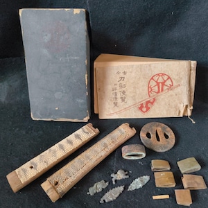 Op de afbeelding: Een assortiment antieke Japanse zwaardfittingen en gerelateerde items. Inclusief een donkergrijs boek, een boekje met Japanse tekst en illustraties, twee houten schede stukken en diverse metalen componenten.
