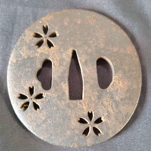 Peut inclure: Une garde d'épée circulaire en métal, appelée tsuba, présentant une patine brune foncée et usée. La surface est ornée de trois motifs de fleurs de cerisier découpés et comporte trois ouvertures centrales pour la lame et les outils.