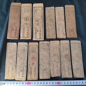 May include: A collection of rectangular, aged paper items with Japanese text and illustrations. The paper has a faded, beige color, and the text appears in black ink. The items are arranged on a surface, with some overlapping. The overall aesthetic is vintage and historical.