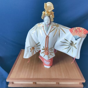 May include: A Japanese doll with a white kimono adorned with gold accents and a red and gold fan. The doll has a serene expression and wears a gold headdress. It stands on a wooden platform.