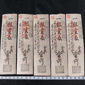 May include: Five antique Japanese paper currency notes. Each note has red and black Japanese characters and a red seal. The notes are rectangular and have a cream-colored background. A ruler is at the bottom of the image.