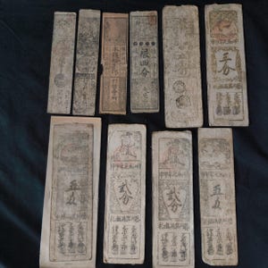 May include: A collection of antique paper currency, featuring various rectangular notes with intricate Chinese characters and illustrations. The notes are aged, with visible wear and tear, suggesting their historical significance. The arrangement is on a dark surface.