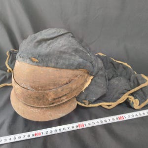 Op de afbeelding: Een antieke helm met een gelaagde, roestige metalen vizier en een donkerblauwe stoffen bekleding. De stof loopt door naar de zijkanten en achterkant, afgezet met een beige rand. De helm is waarschijnlijk uit een historische periode.