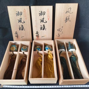 May include: Three rectangular wooden boxes containing pairs of ceramic bells with tassels. The bells feature floral and geometric patterns in blue, gold, and green. The lids are inscribed with Japanese characters and red stamps, measuring 35 cm in length.
