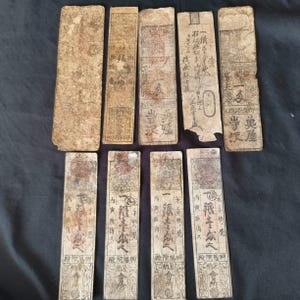 May include: A collection of antique, rectangular paper documents with faded text and red seals. The papers are various sizes and show signs of age, with a worn, beige color. Some have visible text in a script.