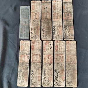 May include: A collection of antique Japanese paper money, featuring rectangular and oblong shapes with faded colors and intricate calligraphy. The bills show signs of age and wear, with visible text and patterns.