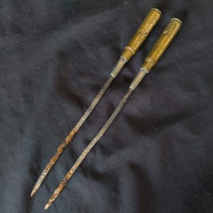 May include: Two antique metal tools with brass handles and slender, rusty shafts. The handles have etched designs. The tools are set against a dark background.