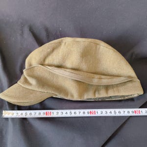 Puede incluir: Sombrero de estilo militar verde oliva con ala curva y una banda alrededor de la corona. El sombrero está hecho de un material similar al fieltro. Una cinta métrica está colocada junto al sombrero.