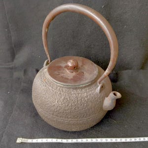 以下が含まれることがあります： 表面がざらざらとした質感の濃い茶色の鋳鉄製ティーポット。高いアーチ型のハンドルと、小さな花形のつまみが付いた蓋が特徴です。下に置かれた白いメジャーは、26センチメートルを示しています。
