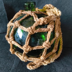 May include: A green glass float encased in a woven tan rope net. The glass has a bottle-like shape with a green neck. The rope net is tightly woven around the glass, creating a decorative nautical item.