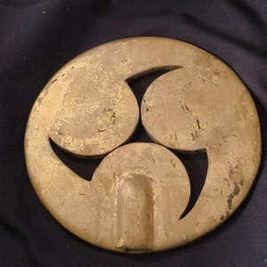 Może przedstawiać: Okrągły, wyglądający na antyczny, złoty metalowy przedmiot z trójczęściowym wzorem. Wzór składa się z trzech zakrzywionych kształtów ułożonych w okrąg, z małym prostokątnym wycięciem pośrodku.