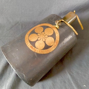 Peut inclure: Une gourde cylindrique gris foncé avec une finition mate et lisse. Un emblème circulaire doré représentant une fleur à six pétales est centré sur le corps. Un cordon de couleur claire est noué autour du goulot pour le transport.