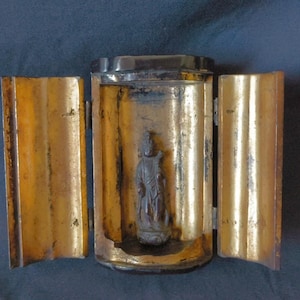 May include: A portable shrine with a dark exterior and a gold-colored interior featuring two hinged doors. Inside the central compartment stands a small, dark, standing figure on a pedestal, enclosed by the gold-finished casing of the shrine.