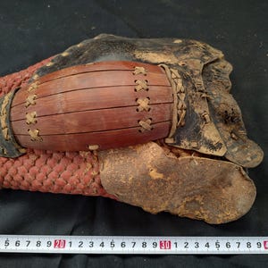May include: Antique Japanese kote, or arm guard, featuring a red woven palm, a dark brown wooden forearm protector, and black leather components. The kote is stitched together with tan leather lacing. The overall design suggests a piece of historical armor.